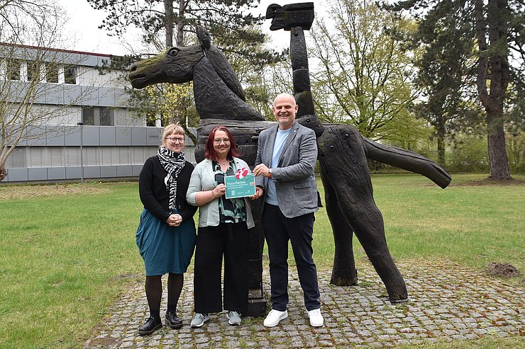 Drei Menschen stehen auf einer Wiese vor einem Holzpferd, es wird eine Plakette übergeben. Ein graues Gebäude steht im Hintergrund. 