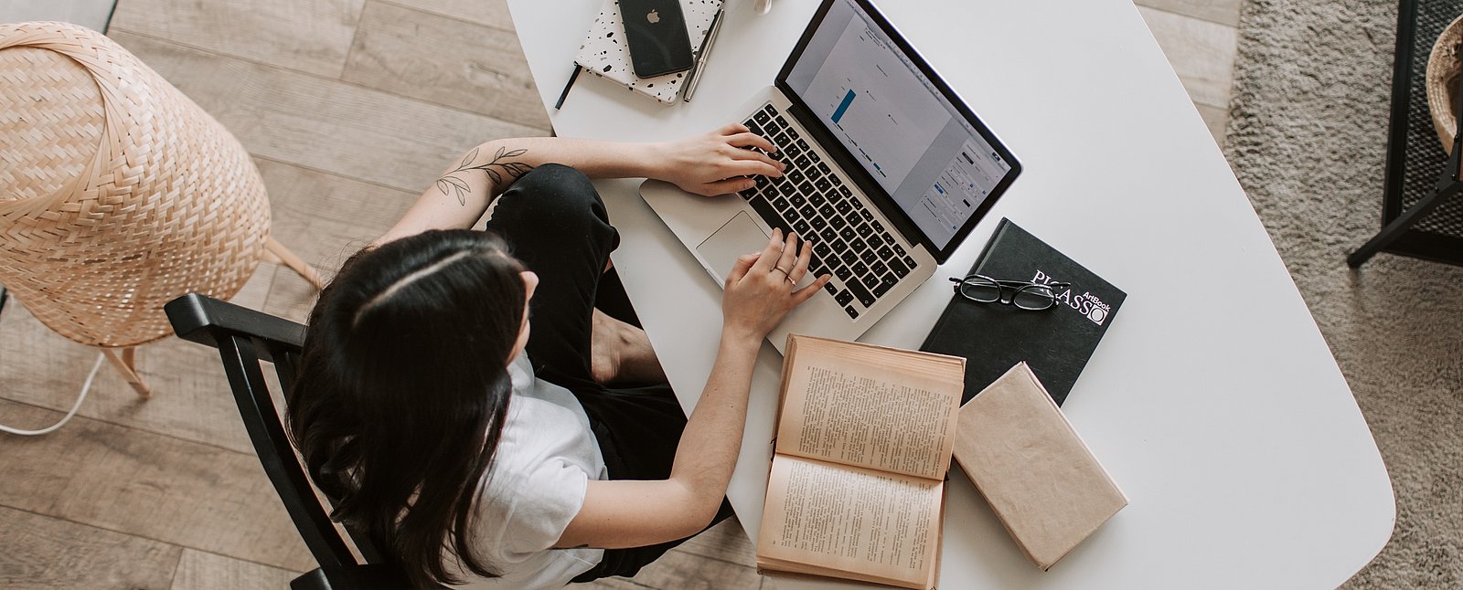 Abgebildet ist eine Frau am Arbeitsplatz mit Notebook und diversen Arbeitsmaterialien