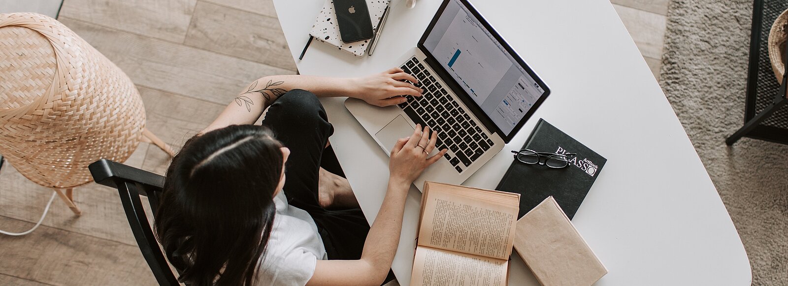 Abgebildet ist eine Frau am Arbeitsplatz mit Notebook und diversen Arbeitsmaterialien